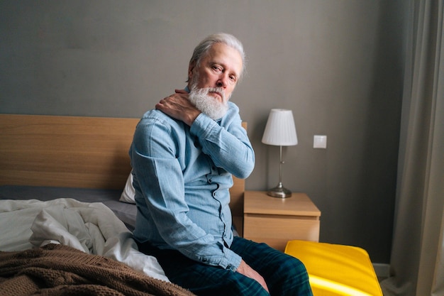 Hombre adulto mayor calentando y masajeando el hombro sentado en la cama por la mañana después de despertarse en casa mirando la cámara