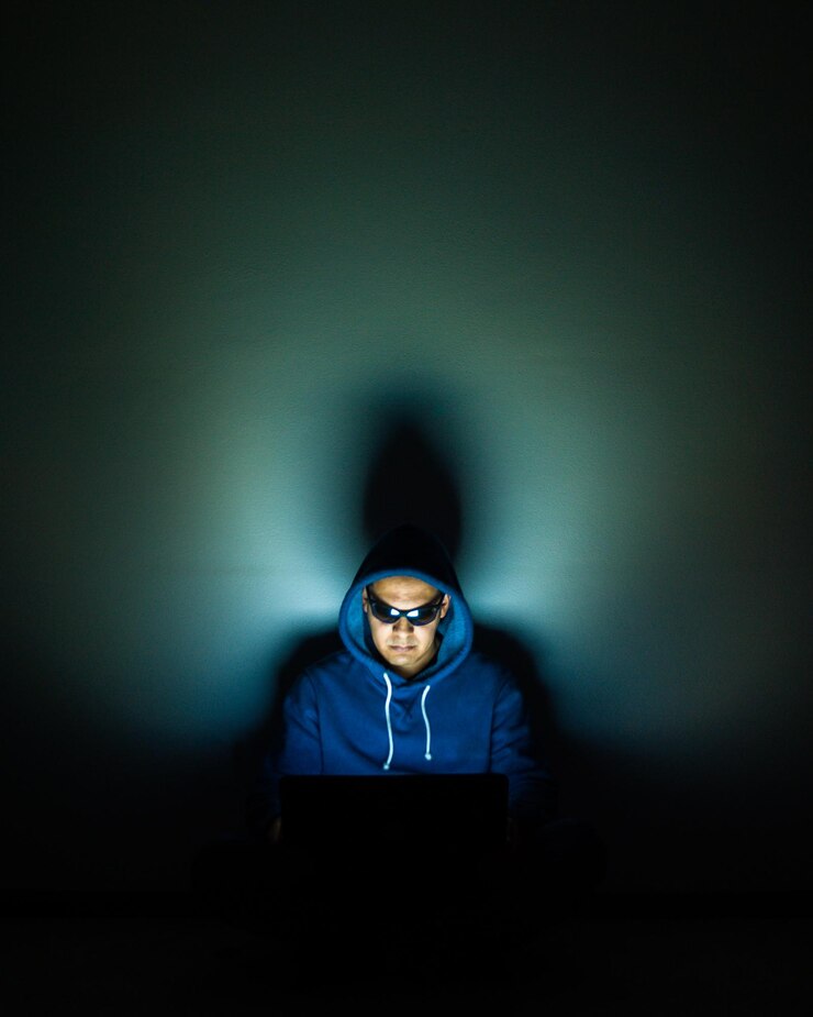 Foto hombre adulto medio con gafas de sol y camisa con capucha mientras usa una computadora portátil contra la pared en el cuarto oscuro
