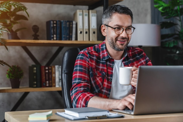 Hombre adulto medio usando una computadora portátil en el escritorio en casa y bebiendo café