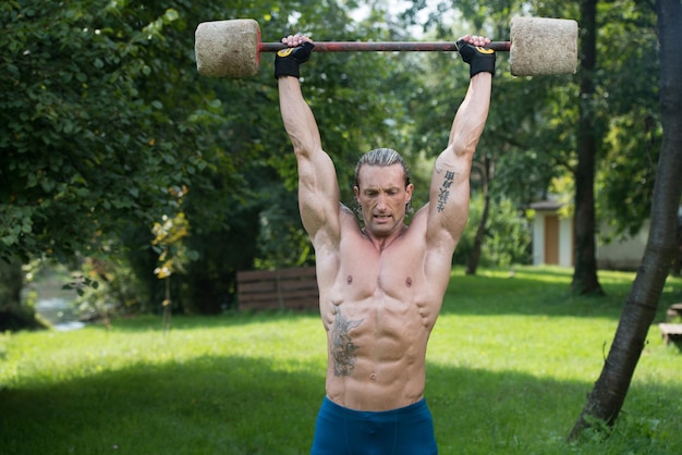 Hombre adulto musculoso haciendo un ejercicio para hombros con entrenamiento al aire libre con barra de mano hecha