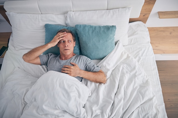 Hombre adulto preocupado acostado en una cama acogedora y mirando la ventana mientras está sumido en pensamientos