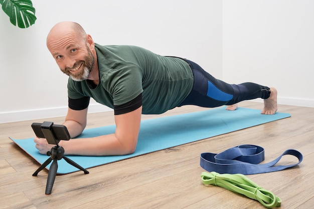 Hombre adulto en ropa deportiva está haciendo ejercicios en la colchoneta frente al teléfono inteligente durante un entrenamiento en línea.