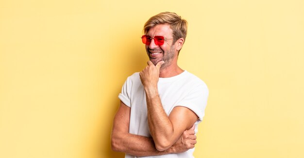 Hombre adulto rubio sonriendo con una expresión feliz y segura con la mano en la barbilla y gafas de sol