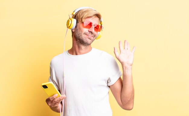 Hombre adulto rubio sonriendo felizmente, saludando con la mano, dándote la bienvenida y saludándote con auriculares