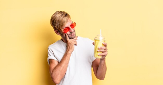 Hombre adulto rubio sonriendo felizmente y soñando despierto o dudando con un batido