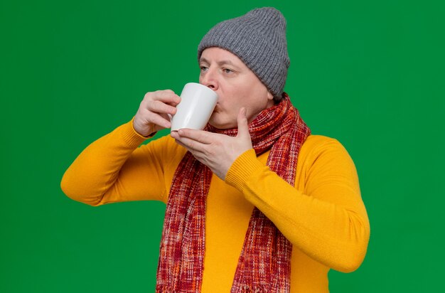 Hombre adulto seguro con gorro de invierno y bufanda alrededor de su cuello bebiendo de taza mirando al lado