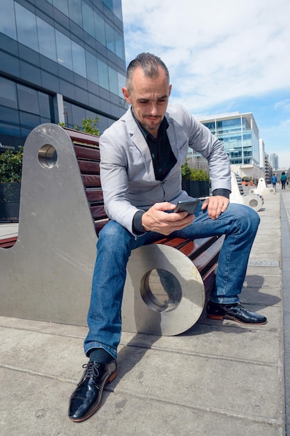 Hombre adulto sentado al aire libre feliz mirando el teléfono revisando las redes sociales y las notificaciones