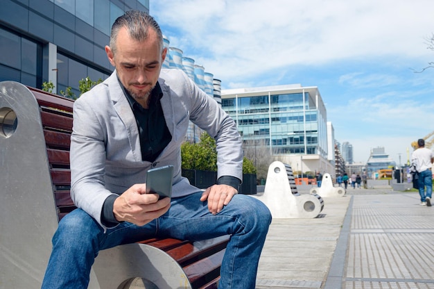 Hombre adulto sentado al aire libre feliz viendo el teléfono revisando redes sociales y notificaciones