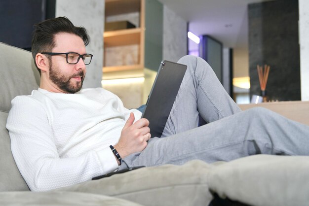 Un hombre adulto serio está usando una tableta en casa acostado en un sofá leyendo un libro electrónico