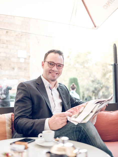 Hombre adulto sonriente en chaqueta con gafas leyendo el periódico mientras está sentado en un café con una taza de café y teléfono y mirando a la cámara por la mañana