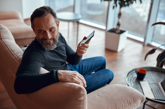 Hombre adulto sonriente sentado en un sillón y sosteniendo un teléfono móvil