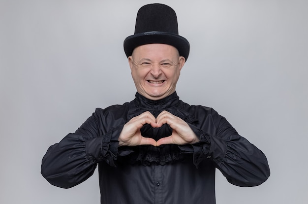 Hombre adulto sonriente con sombrero de copa y camisa gótica negra gesticulando signo de corazón mirando