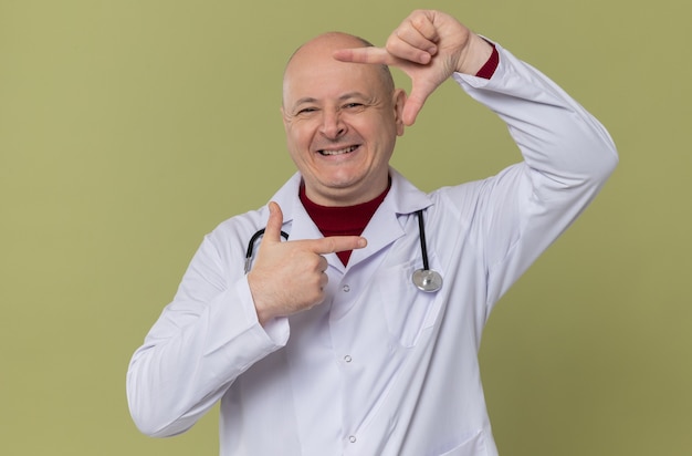 Hombre adulto sonriente en uniforme médico con estetoscopio gesticulando bastidor y mirando