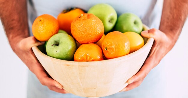 Hombre adulto sosteniendo un balde lleno de frutas de temporada frescas y de colores como orane y manzanas