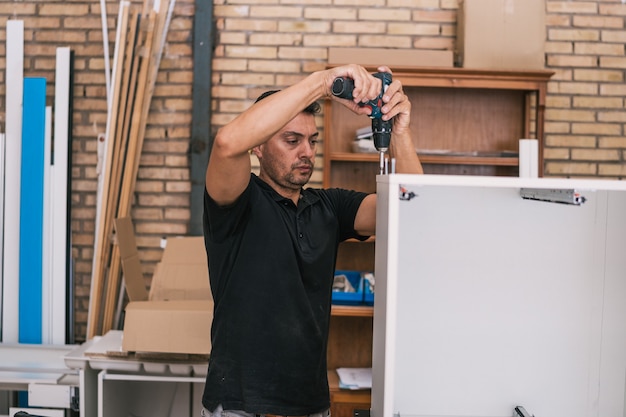 Hombre adulto con un taladro para arreglar dos piezas de una estructura de madera en un taller