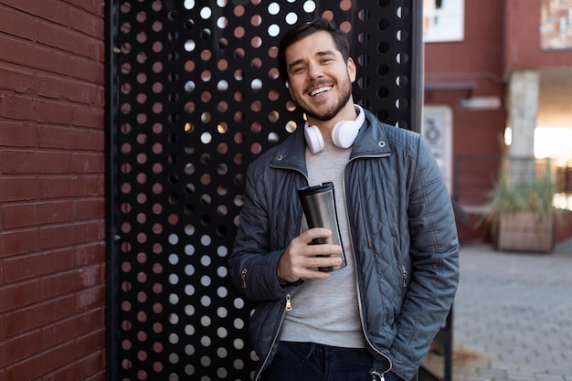 Hombre adulto con una taza de café en las manos y auriculares sonriendo mira la cámara afuera