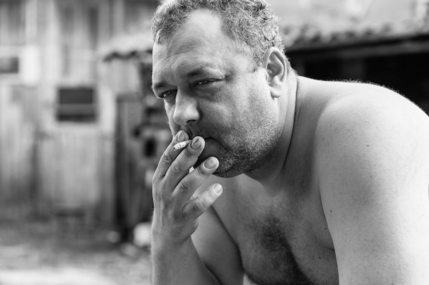 Un hombre adulto con el torso desnudo fuma en el patio de una casa del pueblo. Depresión y estrés. Fotografía en blanco y negro.