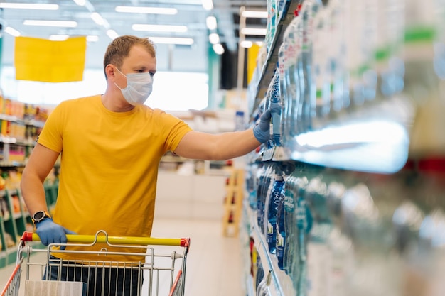 El hombre adulto usa una máscara desechable y guantes de goma como medidas preventivas durante la epidemia elige agua en la tienda minorista trata de estar seguro durante el brote de neumonía por coronavirus Autoaislamiento