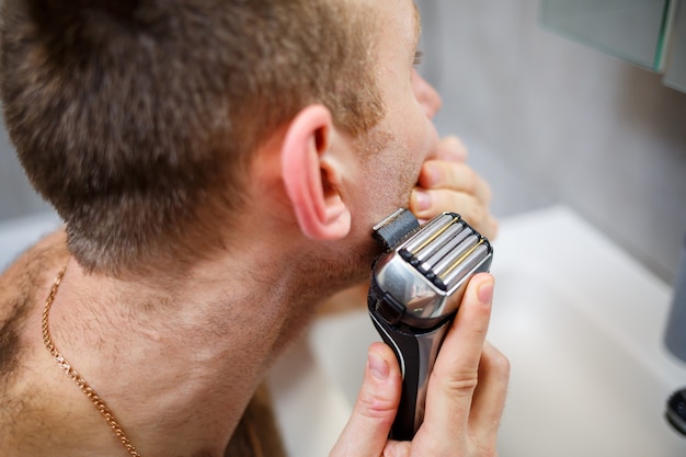 Un hombre se afeita la cara con una maquinilla de afeitar eléctrica frente a un espejo. Irritación de la piel. Procedimiento de baño
