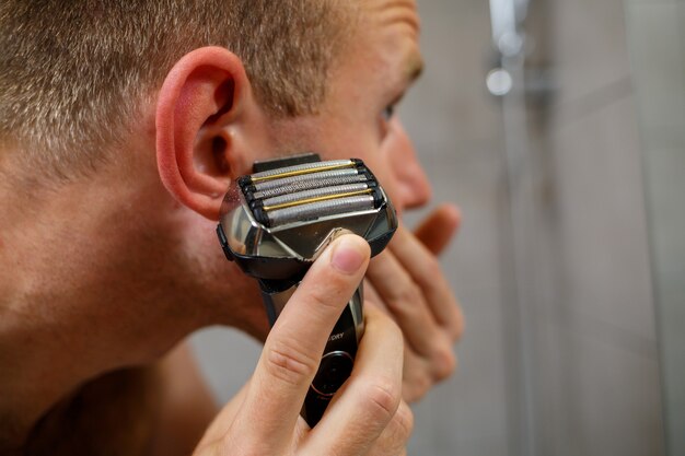 Un hombre se afeita la cara con una maquinilla de afeitar eléctrica frente a un espejo. Irritación de la piel. Procedimiento de baño