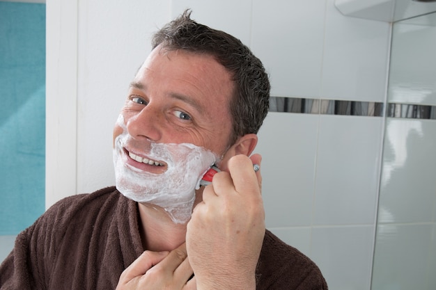 Hombre afeitado con una cuchilla de afeitar y crema de afeitar en el baño.