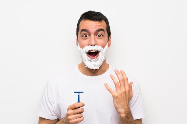 Hombre afeitándose la barba sobre una pared blanca aislada con expresión facial sorpresa