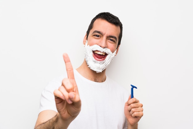 Foto hombre afeitándose la barba sobre una pared blanca aislada mostrando y levantando un dedo