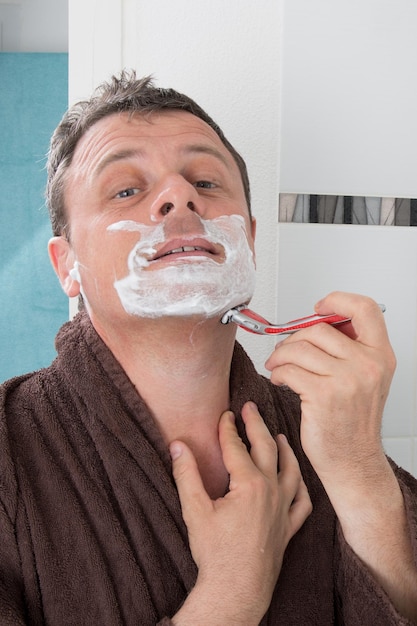 Hombre afeitándose con una hoja de afeitar y crema de afeitar en el baño.