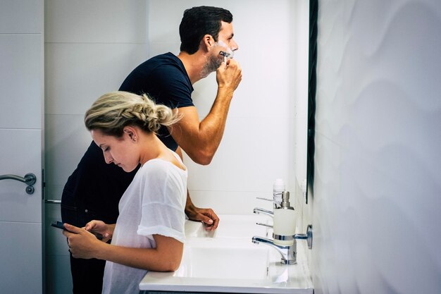 Hombre afeitándose con su novia usando el teléfono en el baño