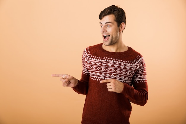 Hombre sin afeitar con cerdas vistiendo un suéter de punto apuntando con el dedo a un lado en copyspace, aislado sobre pared beige