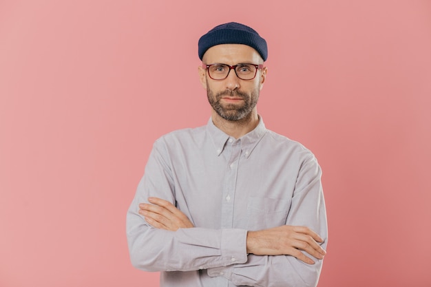 Hombre sin afeitar cruza las manos sobre el pecho, usa gafas y camisa blanca, aislado sobre fondo rosa