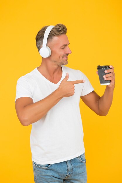 Hombre sin afeitar escucha música y señala con el dedo índice a la taza de café para llevar promocionando y publicitando un fondo amarillo, servicio para llevar.