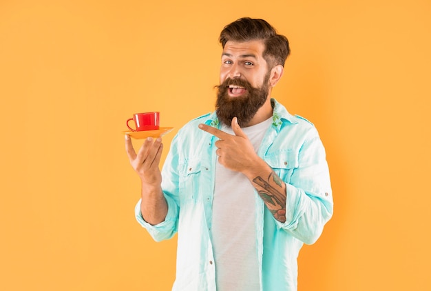 Hombre sin afeitar feliz en camisa señalar con el dedo el café en la taza de café de fondo amarillo