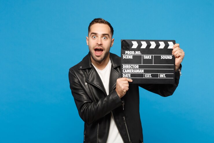 Foto hombre sin afeitar joven elegante hermoso en camiseta blanca de la chaqueta negra sostenga en la mano que hace la película claqueta aislada en el retrato del estudio del fondo de la pared azul. concepto de estilo de vida de personas. simulacros de espacio de copia.