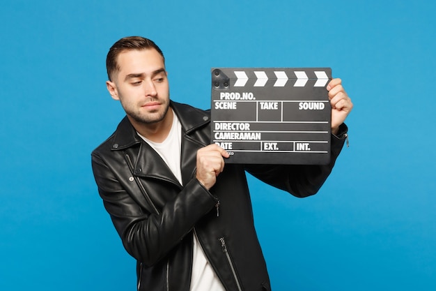 Hombre sin afeitar joven elegante hermoso en camiseta blanca de la chaqueta negra sostenga en la mano que hace la película claqueta aislada en el retrato del estudio del fondo de la pared azul. Concepto de estilo de vida de personas. Simulacros de espacio de copia.