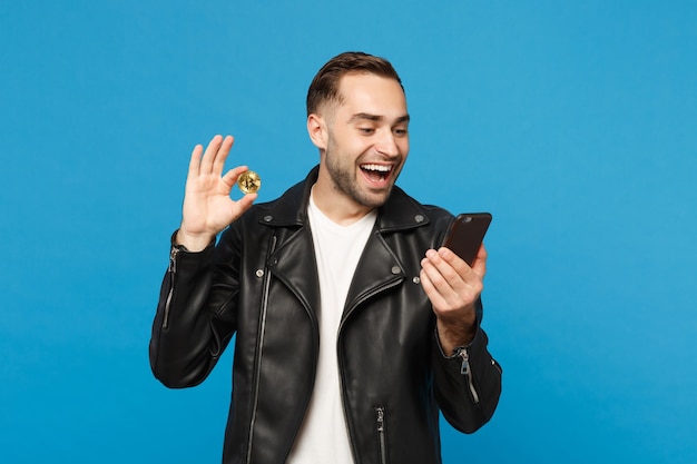 Hombre sin afeitar joven elegante hermoso en la camiseta blanca de la chaqueta negra sostenga en moneda del bitcoin del teléfono móvil de la mano aislada en retrato del estudio del fondo de la pared azul. Concepto de estilo de vida de personas. Simulacros de espacio de copia