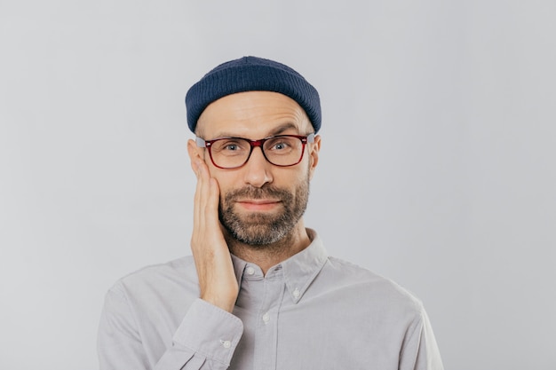 Un hombre sin afeitar de ojos azules levanta las cejas, mantiene la mano en la mejilla, se ve feliz, usa lentes, viste un sombrero negro y una camisa