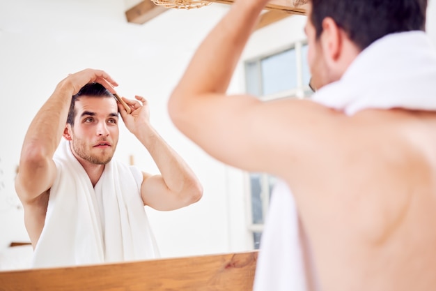 Hombre sin afeitar peinándose mientras está de pie con una toalla blanca en el cuello cerca del espejo en el baño.