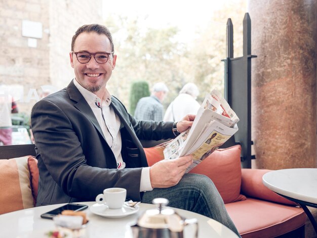 Hombre sin afeitar sonriente en traje negro y gafas leyendo el periódico mientras descansa en un café con una taza de café y un teléfono inteligente y mirando a la cámara