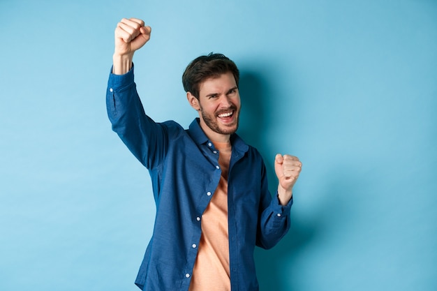 Foto hombre afortunado celebrando la victoria, animando y gritando sí con alegría, levantando la mano como campeón, de pie sobre fondo azul.