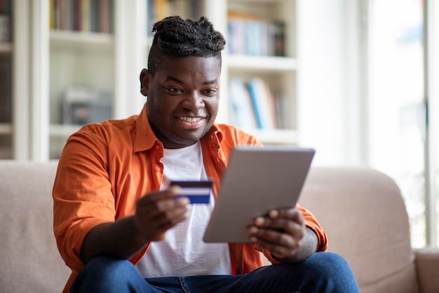 Hombre africano alegre usando libreta digital y tarjeta de crédito