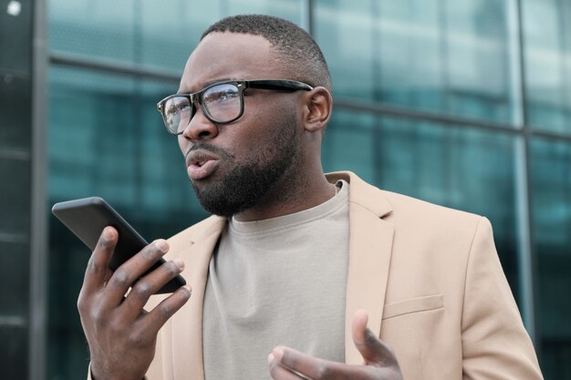 Hombre africano en anteojos grabando un mensaje de audio en el teléfono móvil mientras está de pie al aire libre