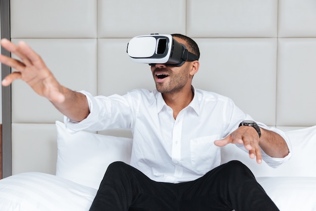 Hombre africano en camisa con dispositivo de realidad virtual sentado en la cama en el hotel