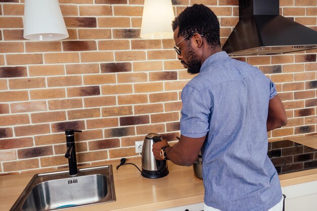 Hombre africano cocinando solo en casa vertiendo sopa en un plato de fondo de campana extractora de pared de ladrillo stewpan