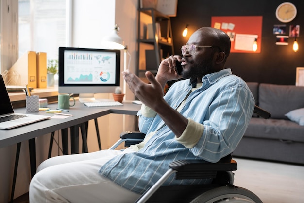 Hombre africano con discapacidad sentado en silla de ruedas y hablando por teléfono móvil, trabaja en casa en la mesa con una computadora portátil