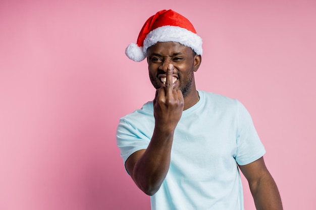 Hombre africano divertido optimista lleno de alegría con cerdas, sonriendo ampliamente, con gorro de Papá Noel en la cabeza, mostrando el dedo medio aislado sobre fondo rosa. Gesto, concepto de emociones.