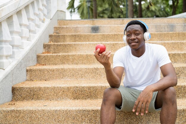 Hombre africano escuchando música con auriculares y sosteniendo manzana roja