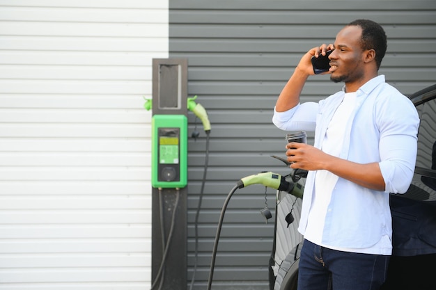 Un hombre africano está parado cerca de un automóvil eléctrico esperando que se cargue en una estación de carga y usando el teléfono