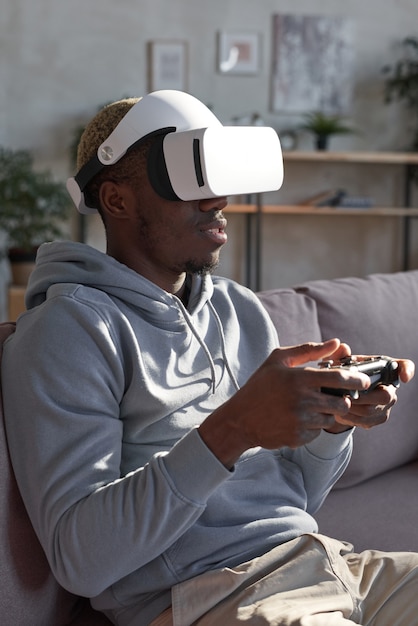 Hombre africano con gafas sentado en el sofá y con joystick que juega videojuegos en casa