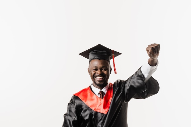 El hombre africano graduado se gradúa de la universidad y celebra los logros académicos Feliz estudiante africano en bata de graduación negra y gorra levanta las manos sobre la cabeza sobre fondo blanco